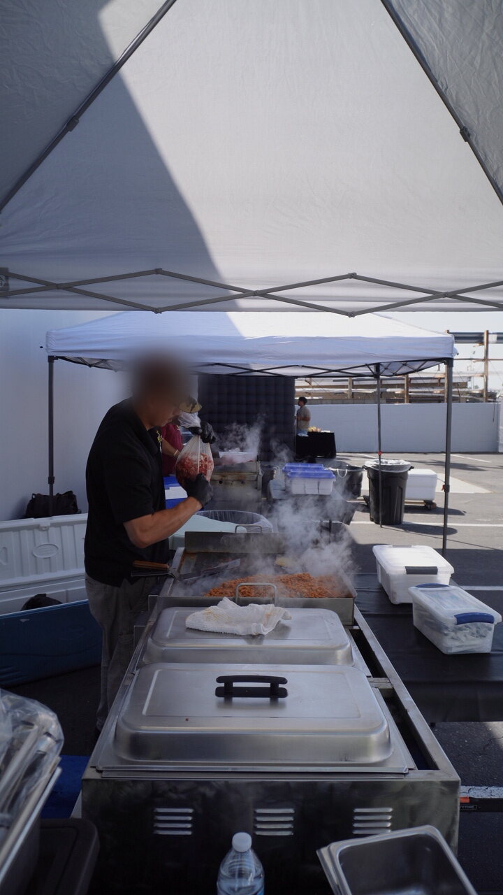 A B&C cook under the canopy at a weekday office lunch in an Anaheim business-park lot, steam rising off the griddle, April 2026