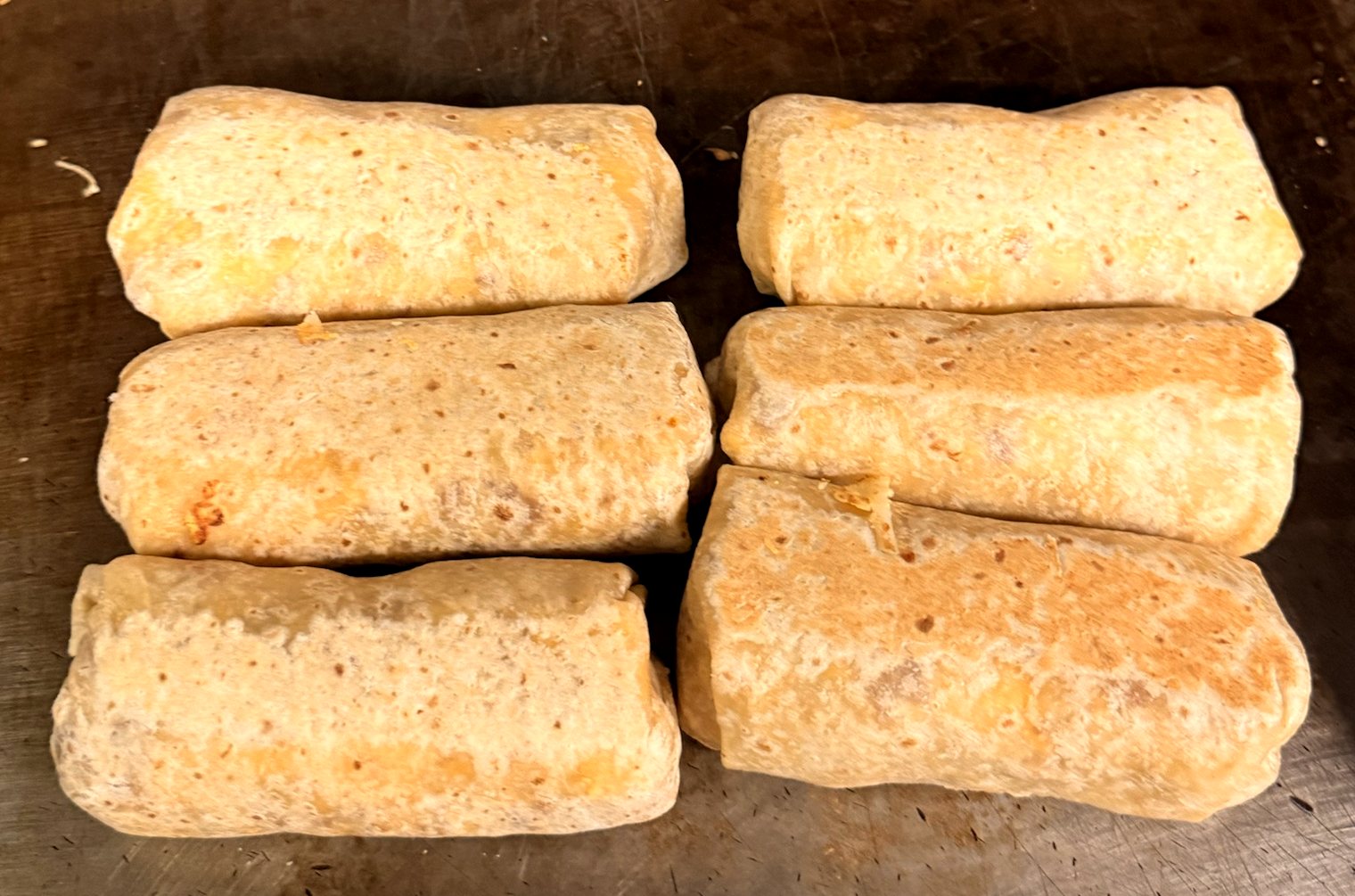 Six wrapped breakfast burritos browning side by side on the B&C griddle