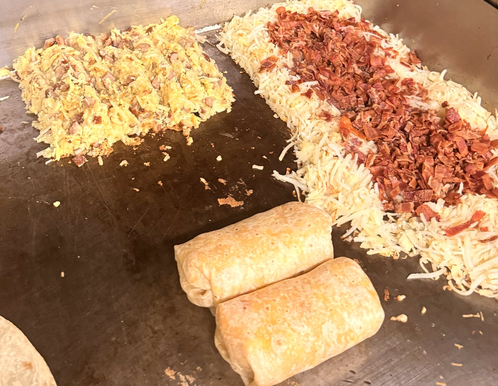 Scrambled eggs, shredded potatoes topped with chopped bacon, and two breakfast burritos on the B&C flat-top griddle