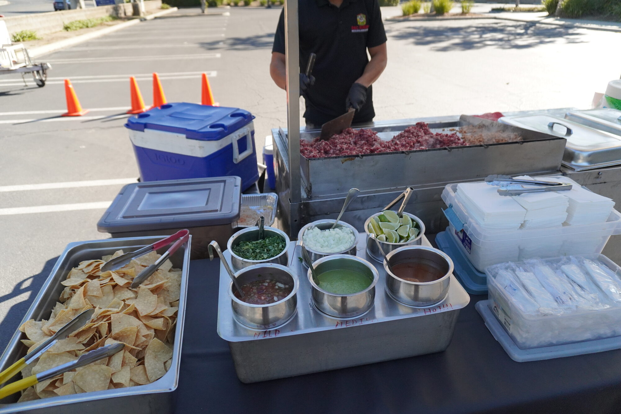 The taco cart mid-service — tortilla chips, three salsas, limes and cilantro along the counter while a cook works the griddle