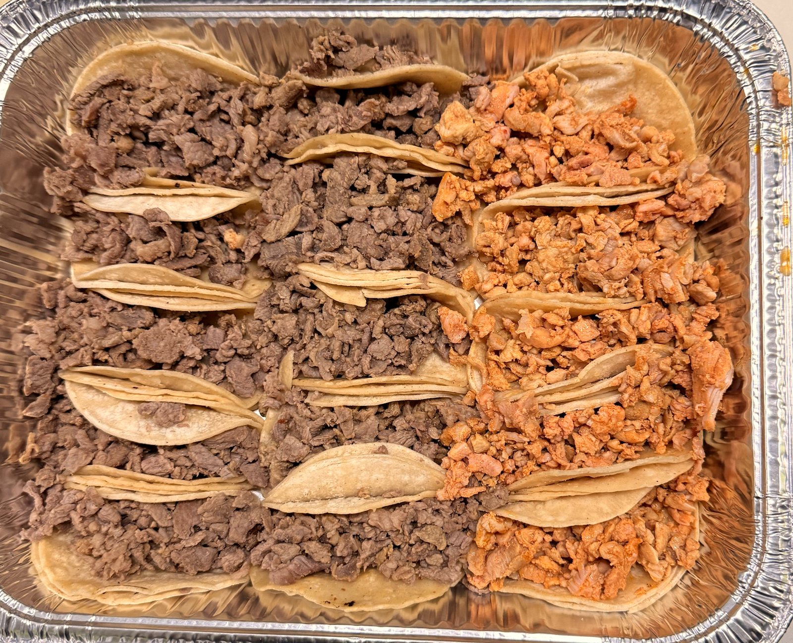 A foil tray of tacos on corn tortillas, two meats side by side