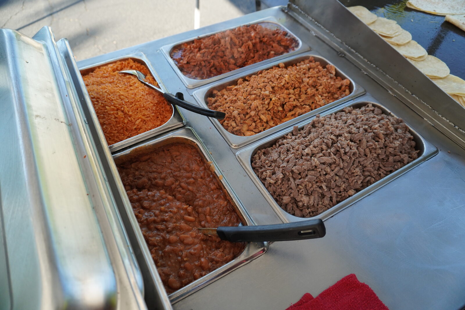 Steel trays of rice, beans and three meats lined up in the cart, tortillas warming behind them