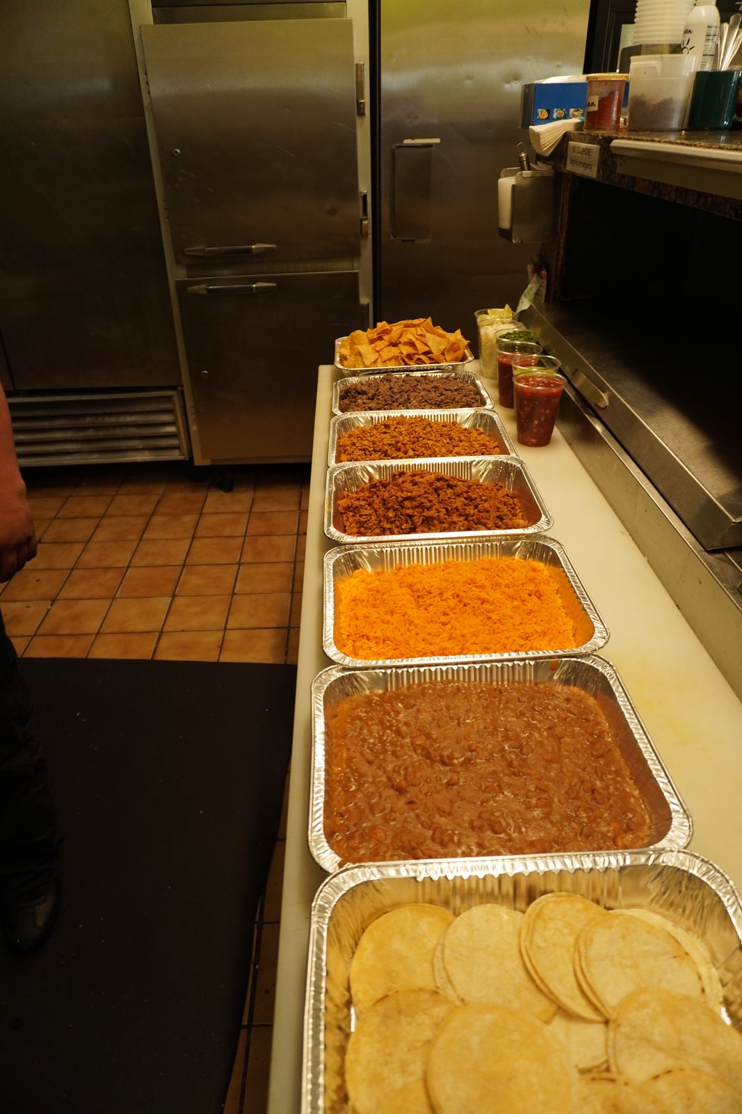 The full pickup order lined up on the pass: foil trays of chips, beans, two meats, Spanish rice, refried beans and warm tortillas