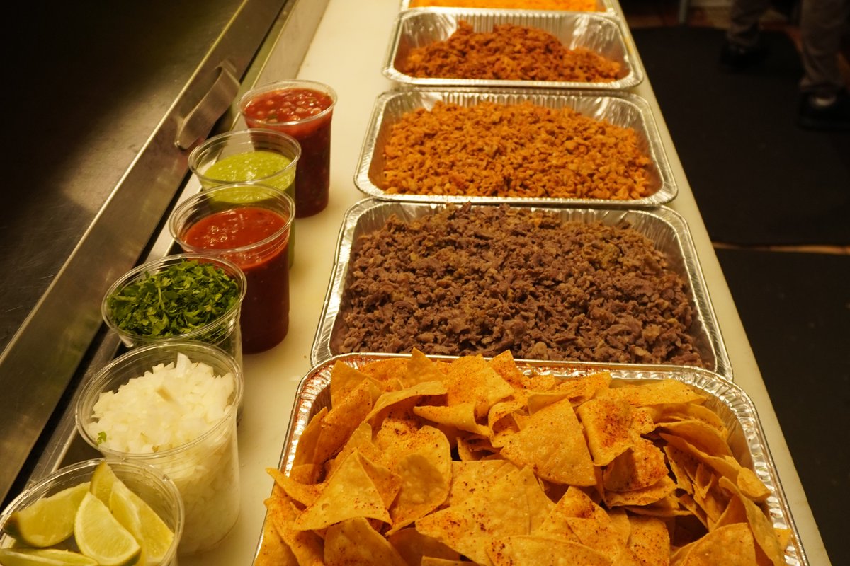 Foil trays of carne asada, Spanish rice, beans and tortilla chips lined up on the pass, with salsas, onion, cilantro and limes cupped alongside