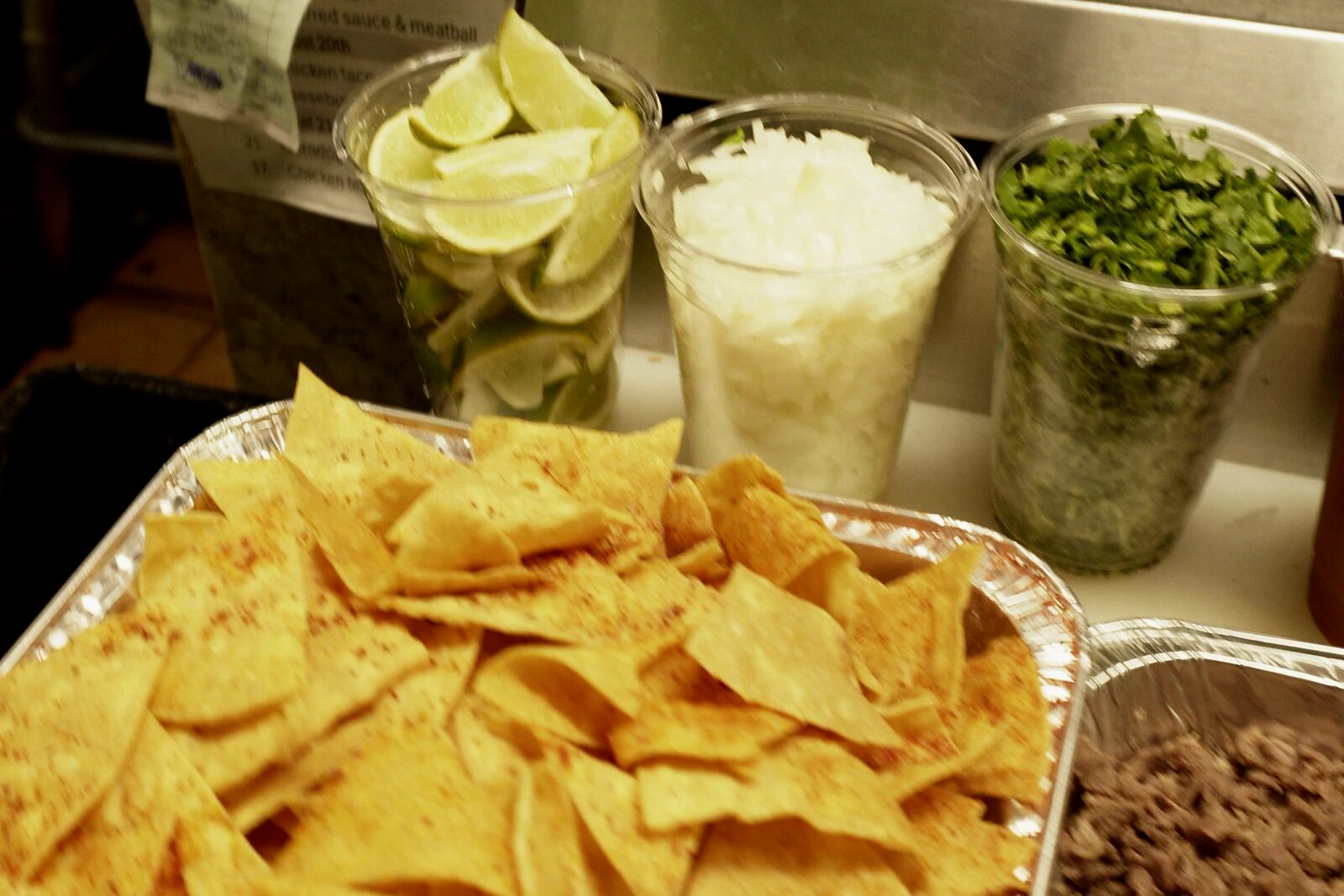 Chips in a foil tray, with limes, onion and cilantro in cups behind